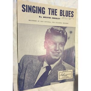 May include: A black and white photo of a man in a suit, singing the blues. The sheet music cover features the title "Singing the Blues" by Melvin Endsley, recorded by Guy Mitchell for Columbia Records. The sheet music is published by Chappell & Co.