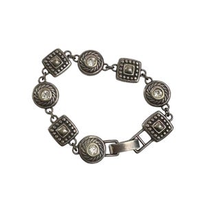 K&ouml;nnte beinhalten: Silberfarbenes Armband mit abwechselnd quadratischen und kreisf&ouml;rmigen Gliedern. Jedes Glied ist mit einem zentralen, klaren Kristall und dekorativen Details verziert. Das Armband hat einen Verschluss.