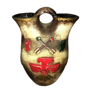 Puede incluir: Jarrón de pared de cerámica vintage con asa y dos aberturas. El jarrón presenta un acabado moteado crema y marrón con ribete dorado. Tiene detalles pintados en rojo, verde y blanco, incluyendo flechas cruzadas y un diseño de pájaro estilizado.