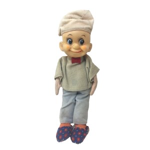 Pode incluir: Uma boneca vintage com rosto pintado, usando um boné bege, uma camisa cinza, calças azuis, uma gravata borboleta vermelha e chinelos estampados. A boneca tem olhos azuis e bochechas rosadas. A boneca está em pé.