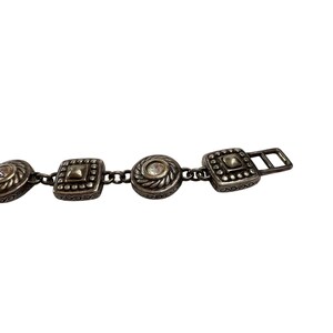 K&ouml;nnte beinhalten: Silberfarbener Metallg&uuml;rtel mit dekorativen quadratischen und runden Verzierungen. Der G&uuml;rtel hat ein Kettendesign und einen rechteckigen Verschluss. Die Verzierungen haben eine strukturierte, antike Oberfl&auml;che und einen kleinen, klaren Stein in der Mitte.