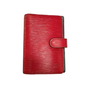 May include: A red leather wallet with a textured surface and a snap closure. The wallet has a rectangular shape and a small, embossed logo on the bottom right corner. The edges are lined with a dark gray material.