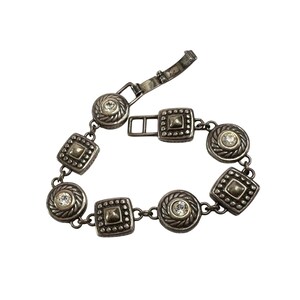 K&ouml;nnte beinhalten: Silberfarbenes Armband mit abwechselnd quadratischen und kreisf&ouml;rmigen Gliedern. Jedes Glied ist mit einem zentralen, klaren Edelstein und einem strukturierten Rand verziert. Das Armband hat einen Verschluss.