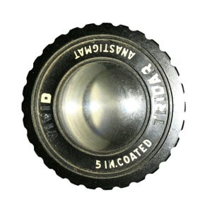 May include: A black camera lens with the text "DIANA ANASTIGMAT TRIOVAR 5 IN. COATED" in white lettering. The lens has a ridged outer edge and a reflective, concave center.