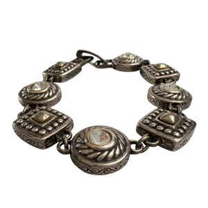 K&ouml;nnte beinhalten: Silberfarbenes Armband mit abwechselnd quadratischen und runden Gliedern. Jedes Glied hat ein zentrales goldfarbenes quadratisches oder rundes Element, umgeben von klaren, runden Steinen. Das Armband hat ein verziertes Vintage-Design.