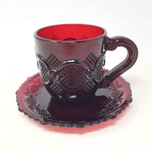 May include: A deep red glass teacup and matching saucer set featuring a raised geometric diamond pattern and circular motifs. The cup has a sturdy handle with decorative detailing, and the saucer has a scalloped edge design.