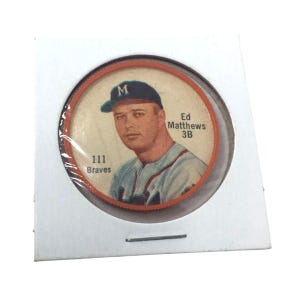 May include: A vintage baseball card featuring Ed Matthews, 3B, of the Braves. The card is circular, set in a protective holder. The card has a portrait of Matthews wearing a baseball cap. The card is in a white holder with a metal staple.