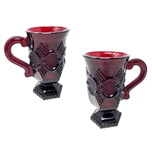 May include: Two ruby red glass mugs with detailed patterns. Each mug has a textured design, a pedestal base, and a curved handle. These mugs are likely for drinks and have a vintage style.