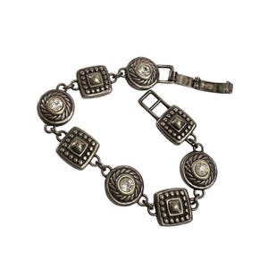 K&ouml;nnte beinhalten: Silberfarbenes Armband mit abwechselnd quadratischen und kreisf&ouml;rmigen Gliedern. Jedes Glied hat einen erhabenen Rand und eine zentrale Verzierung. Die kreisf&ouml;rmigen Glieder haben einen klaren, runden Stein in der Mitte. Das Armband ist ge&ouml;ffnet und zeigt den Verschlussmechanismus.