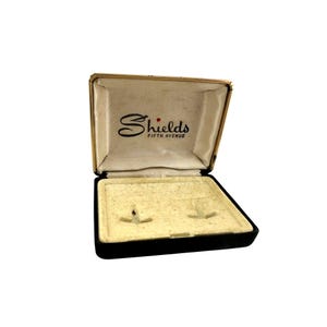 May include: Open vintage jewelry box with a gold-tone frame and black base. The interior is beige with two small slots. The lid features the name "Shields FIFTH AVENUE" in black script.