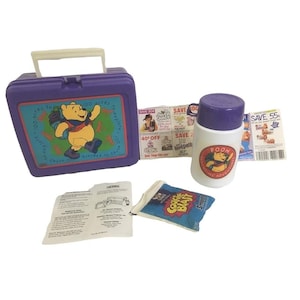 以下が含まれることがあります： ヴィンテージの紫色のランチボックス。くまのプーさんのグラフィック、お揃いの魔法瓶、小さな青いスナックバッグが写っています。ランチボックスには白いハンドルと「100 acres to explore」の文字があります。