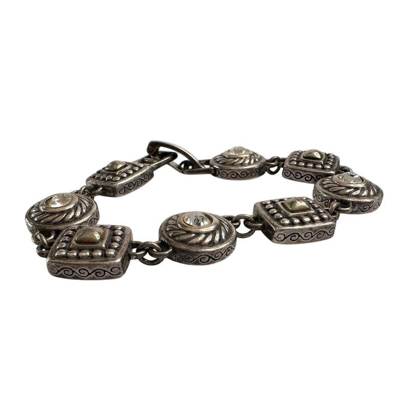 K&ouml;nnte beinhalten: Silberfarbenes Armband mit abwechselnd quadratischen und runden Gliedern. Jedes Glied hat eine goldfarbene Mitte und einen klaren, runden Stein. Das Armband hat ein verziertes, Vintage-Design und einen Verschluss.