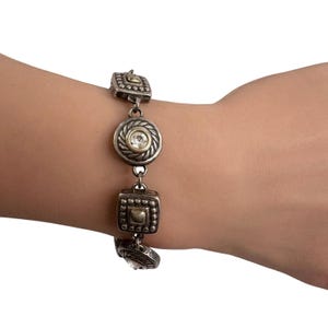 K&ouml;nnte beinhalten: Silberfarbenes Armband mit dekorativen Gliedern. Das Armband hat quadratische und kreisf&ouml;rmige Glieder, jedes mit aufwendigen Details und einem zentralen, klaren Stein. Die kreisf&ouml;rmigen Glieder haben einen goldfarbenen Rand.