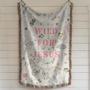May include: A woven throw blanket with a white background and a floral pattern. The text "WILD FOR JESUS" is printed in pink on the blanket.