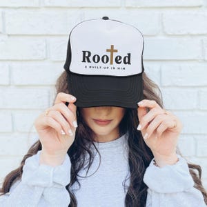 May include: Black and white trucker hat with a gold cross and the text "Rooted & Built Up In Him".