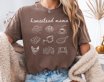May include: A brown t-shirt with the text "homestead mama" in white. The shirt features a series of white illustrations of farm-related items, including a sunrise, a loaf of bread, a cup of coffee, a carton of eggs, a watering can, a frying pan, a chicken, a field of flowers, and a swing set.