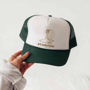 May include: A white and green trucker hat with a brown cowboy boot illustration and the text "STAND FIRM".