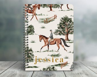 May include: Spiral-bound notebook featuring a watercolor equestrian scene with riders on horseback, trees, and fences. The notebook has a cream-colored background with the name "Jessica" in gold lettering. The notebook is on a wooden surface.