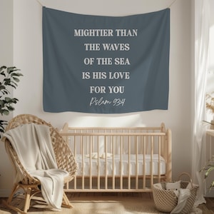May include: A blue wall hanging with white text: "MIGHTIER THAN THE WAVES OF THE SEA IS HIS LOVE FOR YOU Psalm 93:4." It's above a wooden cot and a wicker chair. The room has a neutral color scheme.