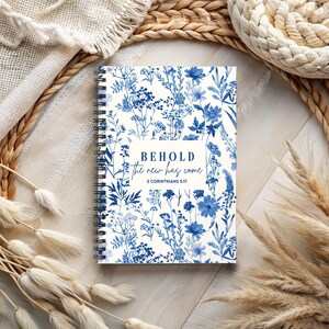 Op de afbeelding: Een wit spiraalnotitieblok met een blauw bloemenpatroon en de tekst "BEHOLD the new has come 2 CORINTHIANS 5:17".