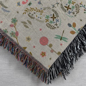 May include: A woven throw blanket with a floral pattern in shades of pink, green, and brown. The blanket has a black fringe border.