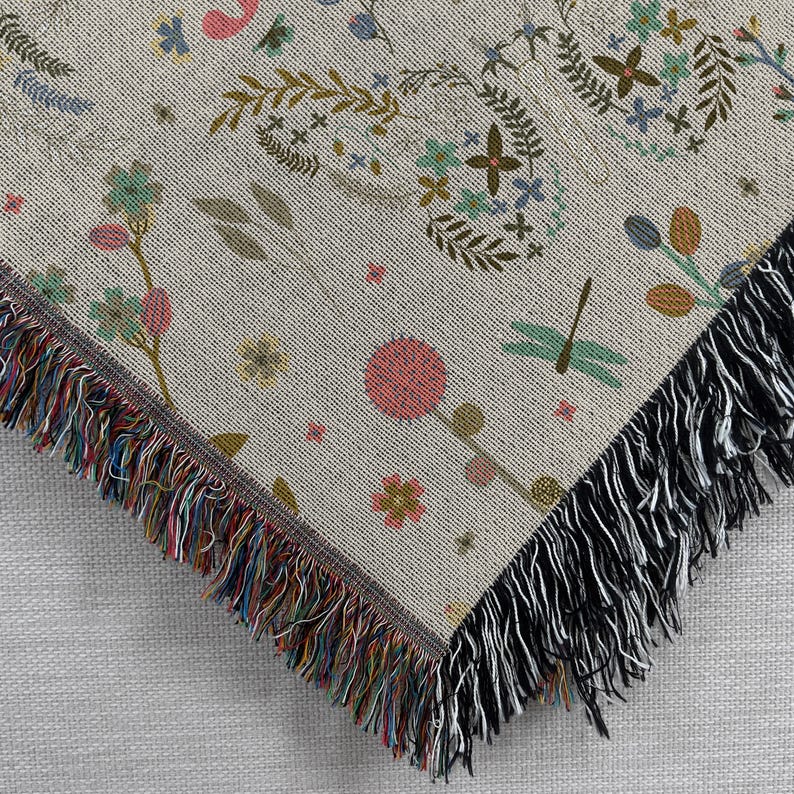 May include: A woven throw blanket with a floral pattern in shades of pink, green, and brown. The blanket has a black fringe border.