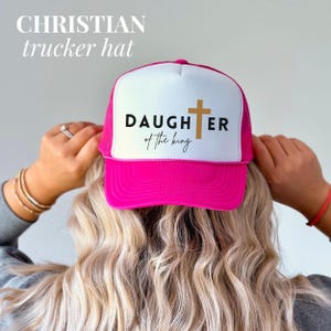 May include: A bright pink and white trucker hat featuring the words "DAUGHTER of the king" and a gold cross. The hat has a mesh back and a curved peak. The words "CHRISTIAN trucker hat" are at the top.