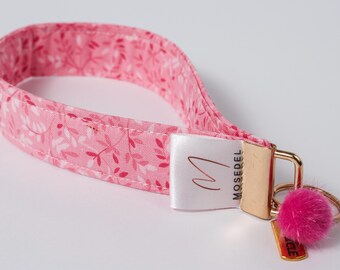 May include: A pink fabric wristlet keychain featuring a white leaf pattern. The strap is secured with a gold-tone metal clasp and a white fabric tag printed with the text MOSEDEL and a logo. A small pink pom-pom hangs from the gold key ring.