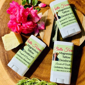 May include: Three white plastic deodorant sticks labeled Bella Forever Magnesium Tallow Deodorant in Peppermint & Tea Tree, Pineapple & Coconut, and Lavender & Vanilla scents, each 0.5 oz, with a block of tallow, pink roses, and rosemary sprigs.