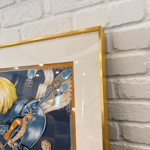 Puede incluir: Un pa&ntilde;uelo de seda cuadrado enmarcado con una ilustraci&oacute;n detallada de equipo ecuestre, que incluye una silla de montar azul, espuelas doradas y plumas amarillas. La obra est&aacute; dentro de un paspart&uacute; blanco ancho y un marco dorado fino.