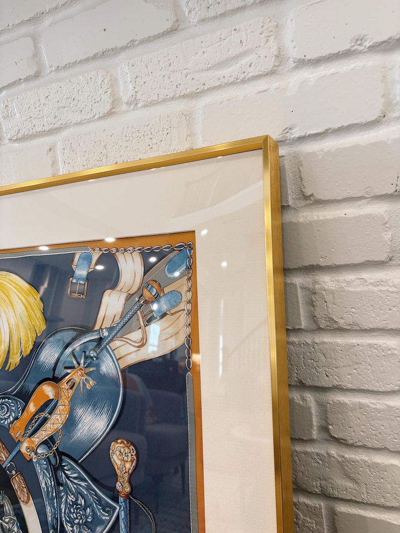 Puede incluir: Un pa&ntilde;uelo de seda cuadrado enmarcado con una ilustraci&oacute;n detallada de equipo ecuestre, que incluye una silla de montar azul, espuelas doradas y plumas amarillas. La obra est&aacute; dentro de un paspart&uacute; blanco ancho y un marco dorado fino.