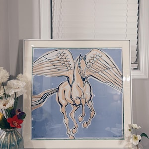 Puede incluir: Una impresi&oacute;n cuadrada enmarcada que presenta una ilustraci&oacute;n estilizada de un caballo alado, o Pegaso, en tonos crema y azul oscuro sobre un fondo azul claro. La obra est&aacute; encerrada en un marco plateado fino con un borde de paspart&uacute; crema.