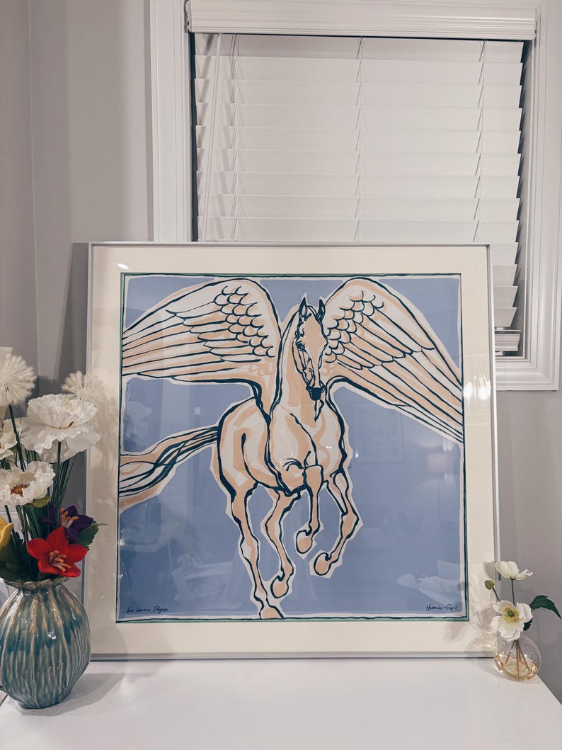 Puede incluir: Una impresi&oacute;n cuadrada enmarcada que presenta una ilustraci&oacute;n estilizada de un caballo alado, o Pegaso, en tonos crema y azul oscuro sobre un fondo azul claro. La obra est&aacute; encerrada en un marco plateado fino con un borde de paspart&uacute; crema.