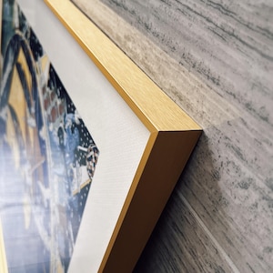 Puede incluir: Una pieza de arte mural rectangular con un marco de metal dorado de acabado cepillado. La obra interior muestra una composici&oacute;n gr&aacute;fica en azul oscuro y amarillo con un borde blanco, protegida por una cubierta de vidrio o acr&iacute;lico transparente.
