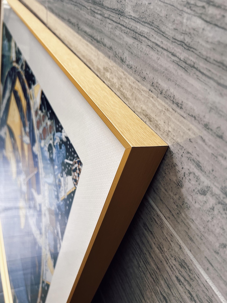 Puede incluir: Una pieza de arte mural rectangular con un marco de metal dorado de acabado cepillado. La obra interior muestra una composici&oacute;n gr&aacute;fica en azul oscuro y amarillo con un borde blanco, protegida por una cubierta de vidrio o acr&iacute;lico transparente.