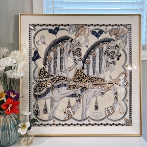 Puede incluir: Una impresi&oacute;n cuadrada enmarcada que muestra dos caballos blancos con crines trenzadas, adornados con arneses negros y dorados con motivos de corazones y plumas. La obra incluye el texto "HARNAIS DE COEUR" y "HERMES PARIS" en la esquina inferior derecha.
