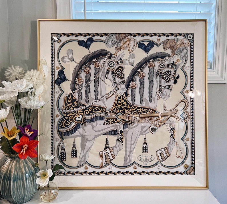 Puede incluir: Una impresi&oacute;n cuadrada enmarcada que muestra dos caballos blancos con crines trenzadas, adornados con arneses negros y dorados con motivos de corazones y plumas. La obra incluye el texto "HARNAIS DE COEUR" y "HERMES PARIS" en la esquina inferior derecha.