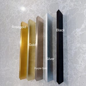 Puede incluir: Cinco tiradores de armario rectangulares dispuestos verticalmente. Los acabados de izquierda a derecha est&aacute;n etiquetados como Rosegold, Gold, Purple Gold, Silver y Black. Cada tirador tiene una textura met&aacute;lica cepillada, excepto el negro mate.