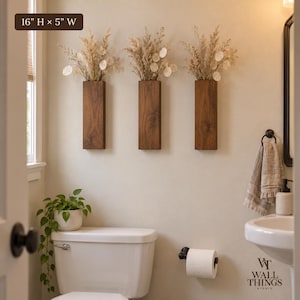 May include: Three rectangular dark wood wall-mounted vases, each measuring 16 inches high by 5 inches wide, containing dried beige grasses and round white seed pods. The vases are arranged in a horizontal row on a light-colored wall.