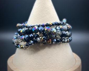 Puede incluir: Un brazalete de múltiples hilos hecho de cuentas negras iridiscentes. El brazalete se muestra en un soporte de madera.