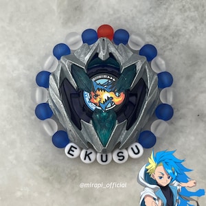 Può includere: Un Beyblade argento e blu con un braccialetto di perline rosse e blu intorno. Il Beyblade ha un design a forma di drago e il testo "Beyblade" su di esso. Il braccialetto ha le lettere "EKUSU" su di esso. L'immagine include anche un personaggio dei cartoni animati con i capelli blu.