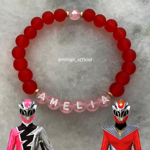Può includere: Braccialetto di perline rosse con un ciondolo di cristallo rosa e il nome "AMELIA" scritto in perline rosa. Il braccialetto è affiancato da due action figure che indossano costumi da ranger rossi e rosa.