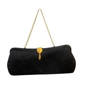 Könnte beinhalten: Eine schwarze Clutch-Tasche mit einem goldfarbenen Kettenriemen. Die Tasche hat einen goldenen Verschluss mit Quastendetail. Die rechteckige Tasche scheint aus einem weichen Stoff zu sein.