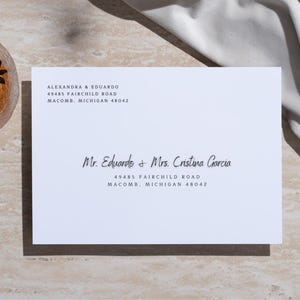 May include: White envelope with a black address label. The address reads: Alexandra & Eduardo, 49485 Fairchild Road, Macomb, Michigan 48042. Below the address is another black address label that reads: Mr. Eduardo & Mrs. Cristina Garcia, 49485 Fairchild Road, Macomb, Michigan 48042.