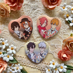 May include: Heart-shaped anime character badges. Each badge features a different pair of characters, forming a heart shape with their hands. The badges are peach-colored with detailed illustrations and surrounded by floral decorations.