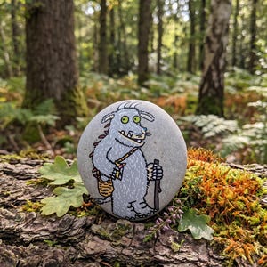 May include: A smooth, round grey stone featuring a hand-painted illustration of a furry, light blue creature with large yellow eyes, a wide grin, and sharp teeth. The creature carries a brown walking stick and a striped satchel over its shoulder.