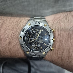 May include: A silver wristwatch with a black face and gold detailing. The watch has several sub-dials and a numbered bezel. The watch is marked "EPSILON ALBA TITANIUM".