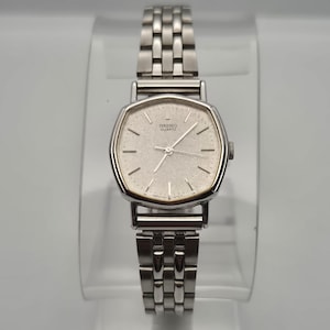 May include: A silver Seiko Quartz watch with a rectangular face and a linked metal strap. The watch face is a light silver colour with black hour markers and hands. The watch is displayed on a clear stand.