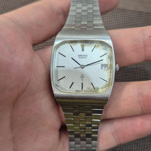 Op de afbeelding: Een vintage Seiko Quartz horloge met een zilverkleurige rechthoekige kast en een bijpassende schakelarmband. De wijzerplaat is zilverkleurig met zwarte markeringen en wijzers, en een datumweergave op 3 uur. Een klassiek horloge.