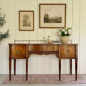 May include: A wooden sideboard featuring a central drawer flanked by two cabinets, each with circular brass pulls. The piece has fluted detailing, tapered legs, and a brass gallery rail along the top edge, displaying a natural wood grain finish.
